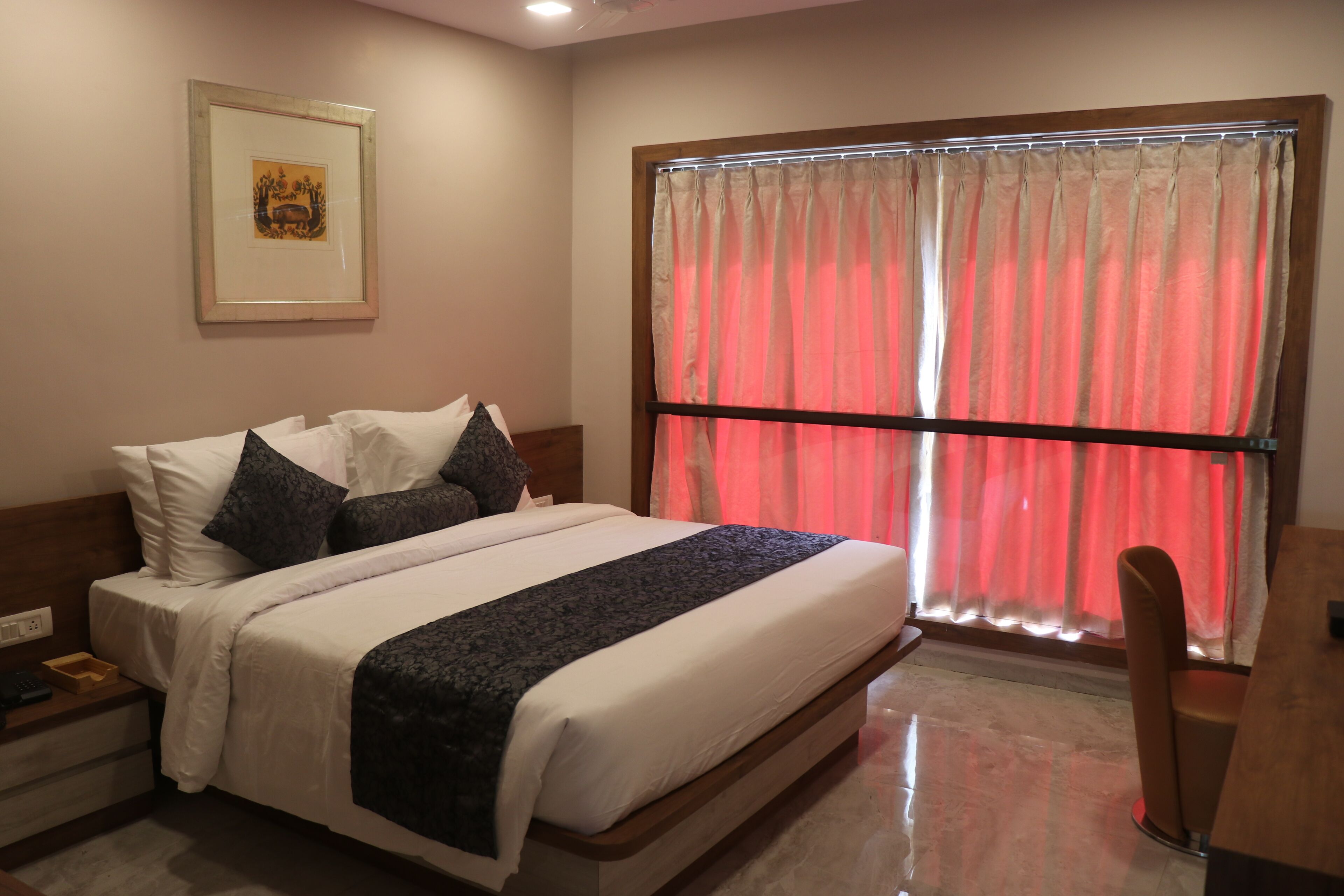 A neatly made bed with white and dark bedding, a framed picture on the wall, and a window with red curtains.