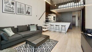 A modern living room with a grey sofa, a coffee table, and a patterned rug. There is a kitchen area with a bar and stools, and a staircase leading to an upper level. - Chalet Park by Maier Mäder (Mäder)