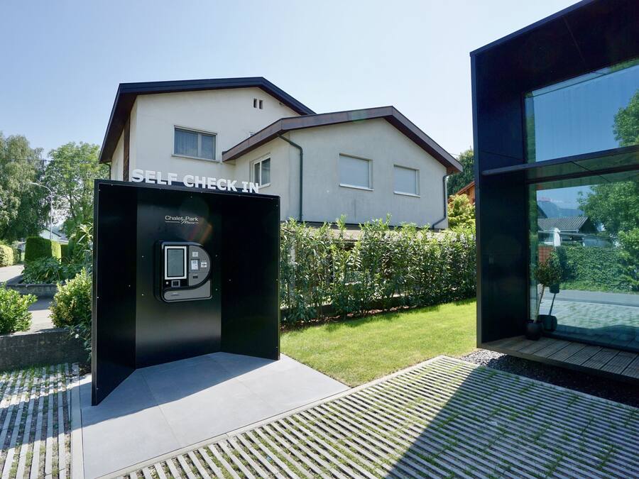 Un quiosco de auto-registro con pantalla digital, ubicado frente a una casa moderna con jardín visible y cielo despejado.