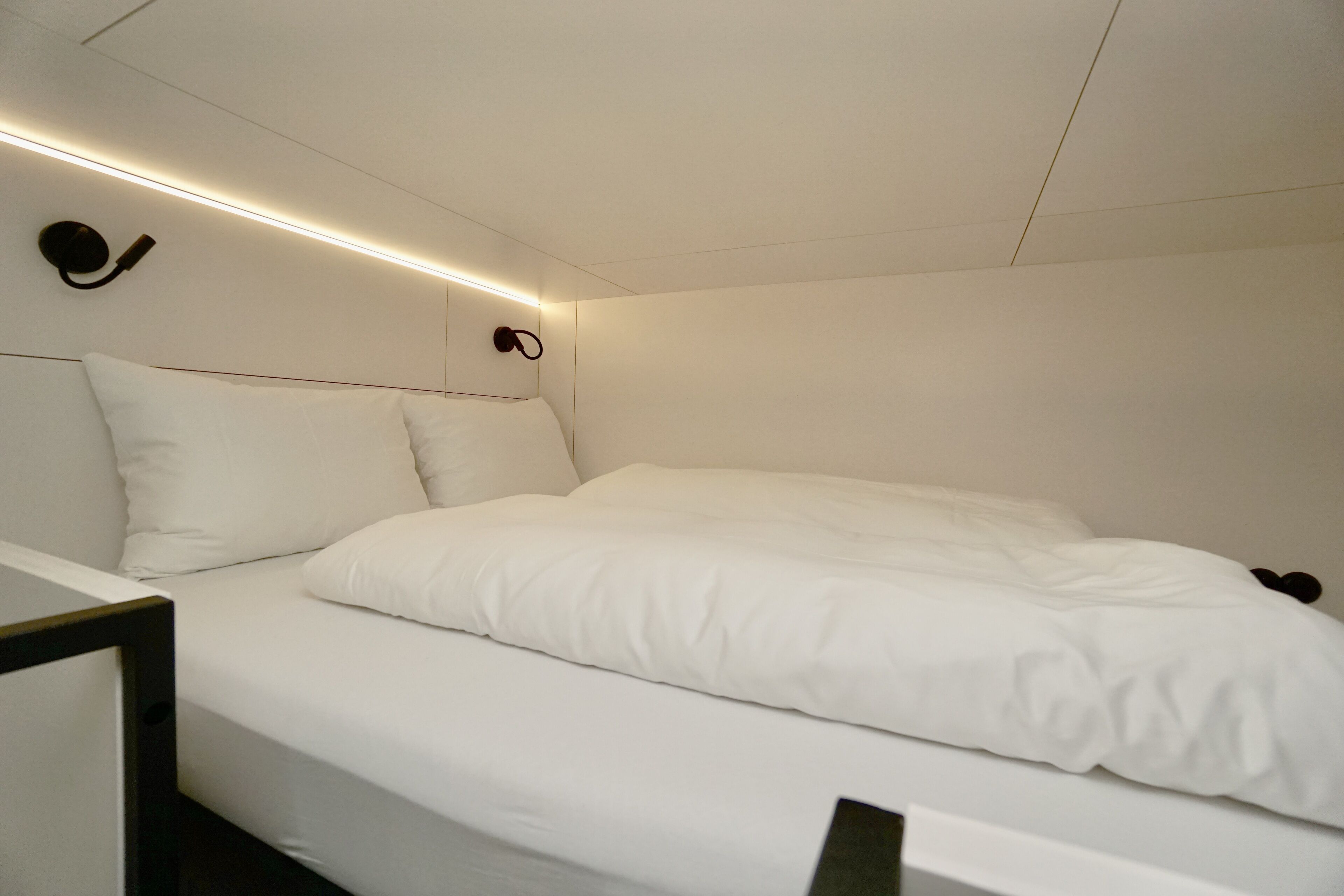 Une chambre à coucher moderne et minimaliste, avec un lit soigneusement fait, une tête de lit dotée d’un éclairage intégré et une décoration aux couleurs sobres et blanches.