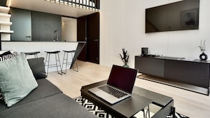 A modern living room with a sofa, a coffee table with a laptop, a kitchen area with a bar stool, and a large TV mounted on the wall. - Chalet Park by Maier Mäder (Mäder)