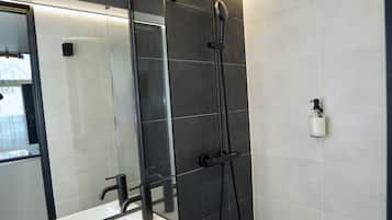 Une salle de bain moderne avec un grand miroir, un lavabo blanc, une pomme de douche noire et un flacon de savon fixé au mur.