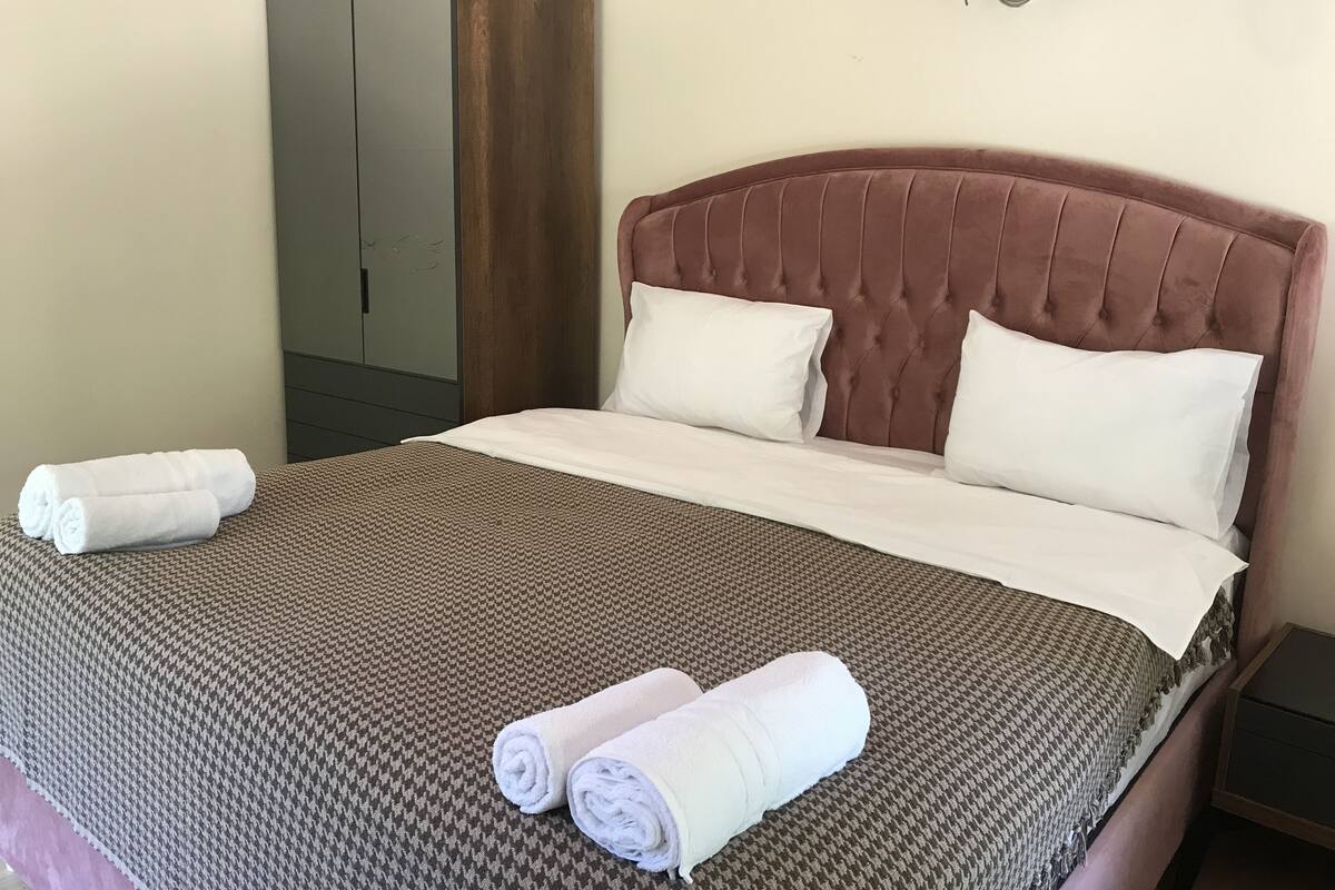 A neatly made bed with two pillows and two rolled towels, a brown houndstooth bedspread, and a wooden wardrobe.