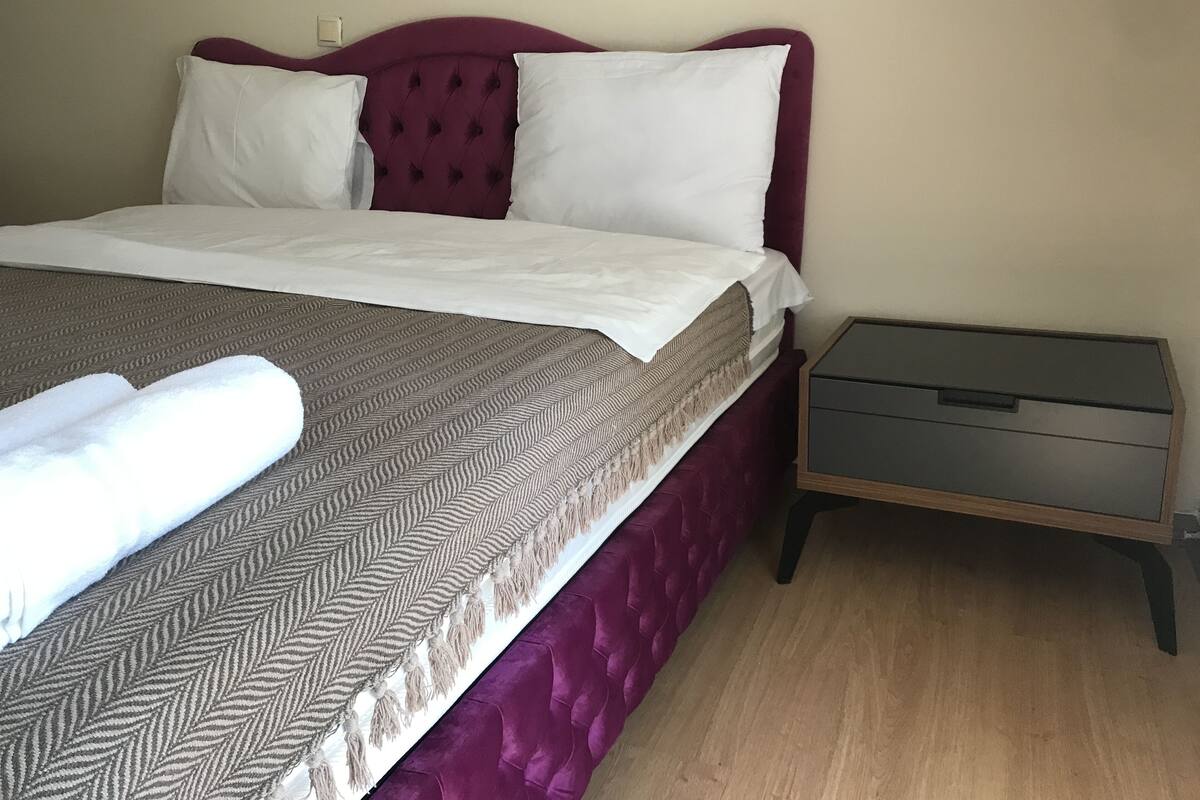 A neatly made bed with a patterned bedspread and a white towel, a wooden nightstand with a drawer, and a plain wall.
