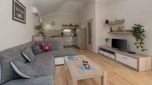Un salon moderne avec un canapé gris, une table basse en bois et un téléviseur posé sur un meuble en bois. On aperçoit, au fond, une cuisine avec un dosseret carrelé et des placards en bois.