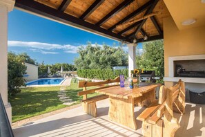 Ein überdachter Außenbereich mit einer Holzbank, einem Grill und einem Pool im Hintergrund.