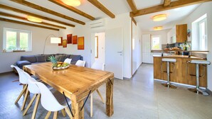 Eine moderne Küche mit Holzbalken, ein Essbereich mit Holztisch und weißen Stühlen sowie eine Bar mit Hockern.