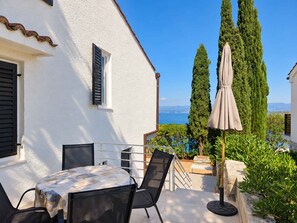 A patio with a table and chairs, a view of the sea, and a tall cypress tree.