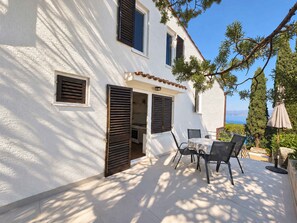 Un edificio blanco con varias ventanas y persianas, un patio con mesa y sillas, y vista al mar.
