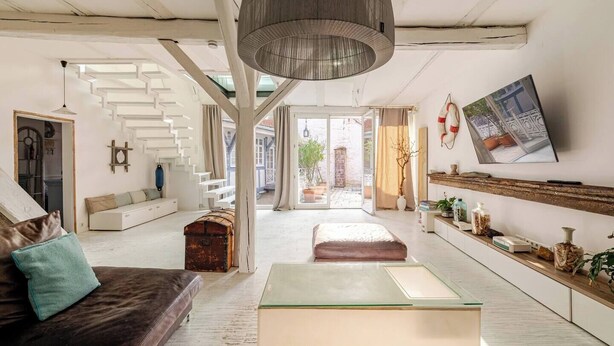 A spacious living area with a staircase, a large pendant light, a sofa with cushions, a glass coffee table, and a TV mounted on the wall.
