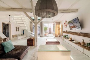 A spacious living area with a staircase, a large pendant light, a sofa with cushions, a glass coffee table, and a TV mounted on the wall.