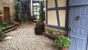 A cobblestone courtyard with potted plants, a wooden door, and a window with a view of the street.