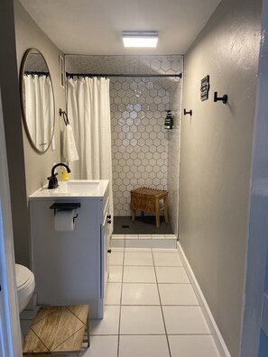 Ein Badezimmer mit weißem Waschbecken, Spiegel und Toilette. Es gibt einen Duschbereich mit sechseckischen Fliesen und einen Holzschemel.