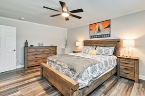 Una camera da letto con un letto in legno, una ventola a soffitto, un comodino con lampada, una cassettiera e un quadro appeso al muro.