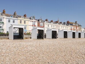 A row of beach huts with open doors, set on a pebbled beach, with colorful buildings in the background. - Royal Terrace View (Weymouth)