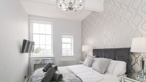 A modern bedroom with a large bed, two bedside lamps, a TV, and a view of the city through the windows.
