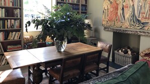 Una habitación con una mesa de comedor de madera, sillas, una estantería, una planta grande en maceta, una lámpara y un tapiz en la pared.