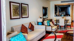 Un salon moderne avec un canapé, des fauteuils, une table basse et un tapis. On aperçoit un coin cuisine à l’arrière-plan.