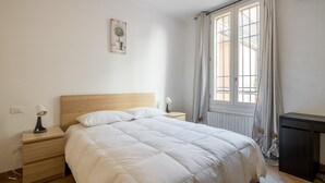 Ein Schlafzimmer mit einem hölzernen Kopfteil, einer weißen Bettdecke, einem Nachttisch mit Lampe und einem Fenster mit Vorhängen.