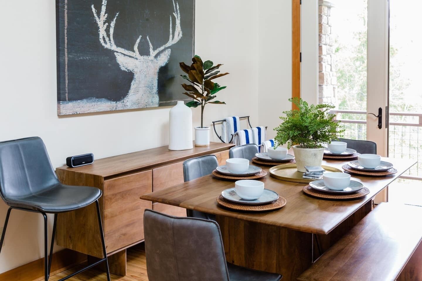 Comedor con mesa de madera dispuesta para cuatro, rodeada de sillas, un cuadro grande de un ciervo y una planta en maceta.