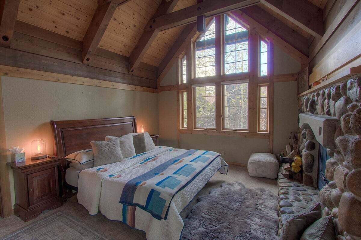 Un dormitorio con techo de madera, una cama grande con colcha, una pared de piedra y una ventana de varios cristales.