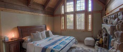 Un dormitorio con techo de madera, una cama grande con colcha, una pared de piedra y una ventana de varios cristales.