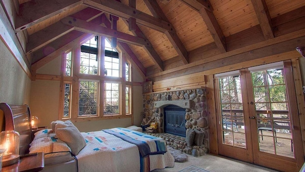 A cozy bedroom with a stone fireplace, wooden beams, and large windows.