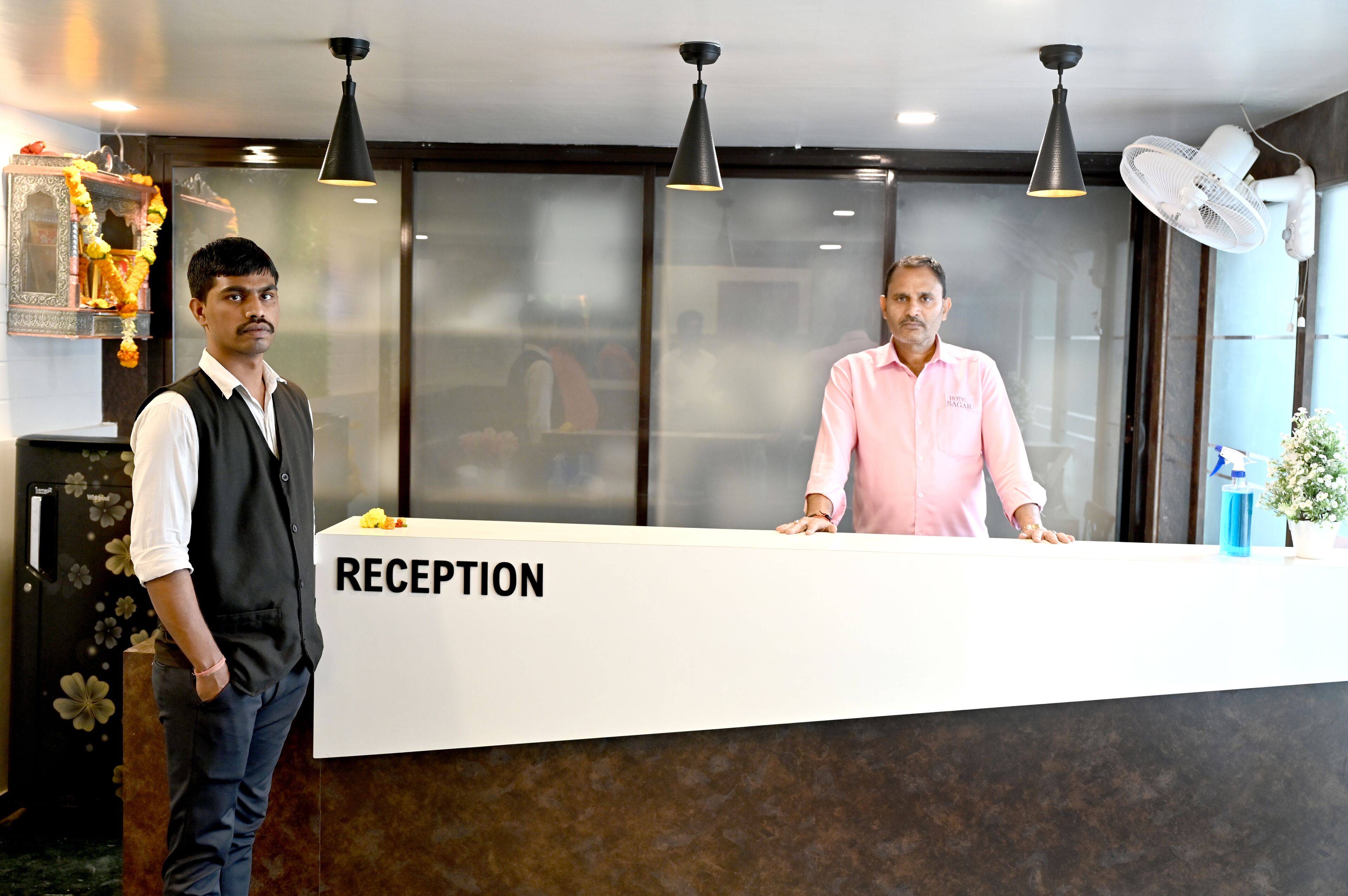 A reception desk with a sign that reads "RECEPTION". There are two people standing behind the desk, one wearing a black vest and white shirt, and the other in a pink shirt.