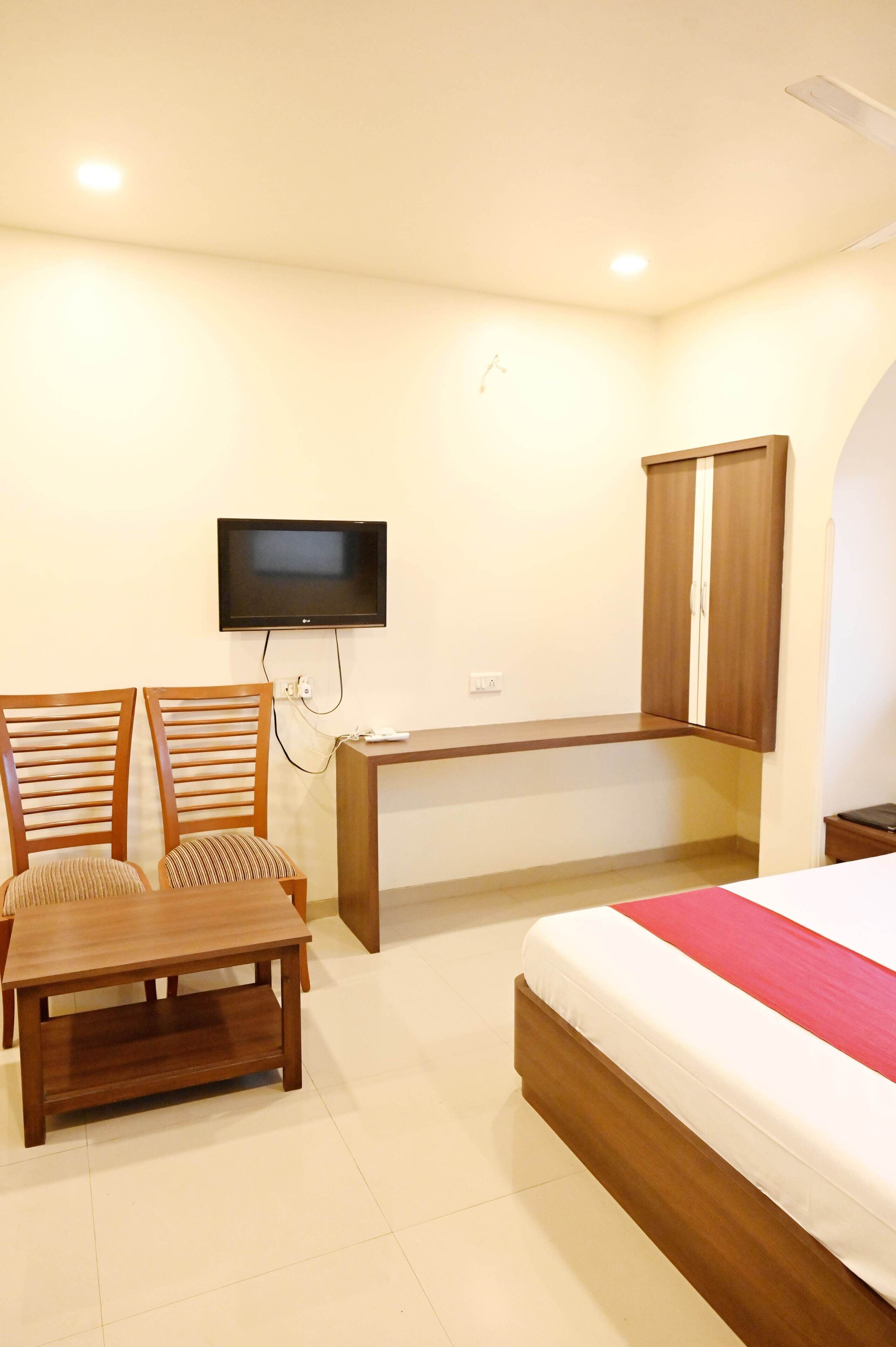 A bedroom with a bed, a TV mounted on the wall, a wooden desk, two chairs, and a wardrobe.