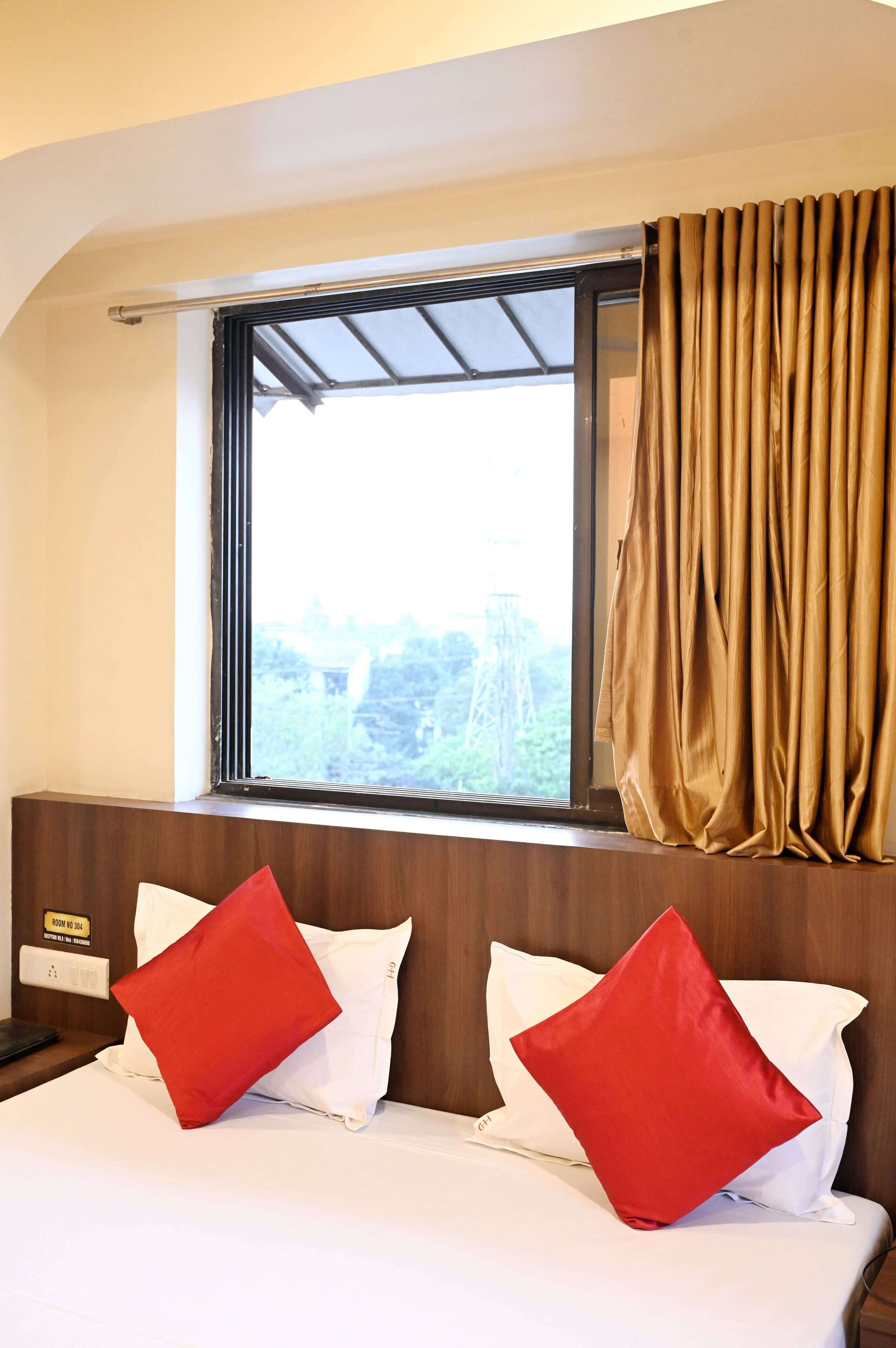 A hotel room with a bed, two red pillows, a window with a view of greenery, and a curtain.