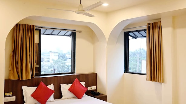 Habitación de hotel con cama, dos ventanas con cortinas, ventilador de techo y vistas al exterior.