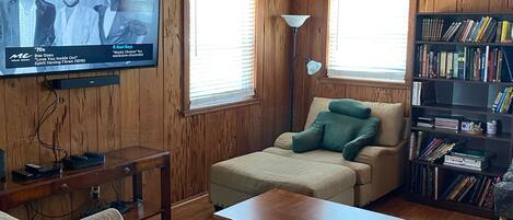 A cozy room with a wooden ceiling, a television mounted on the wall, a sofa, a coffee table, a bookshelf, and a window with blinds.