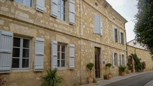 A two-story building with white shutters, a central entrance, and potted plants along the sidewalk. - LA VILLA (Terraube)