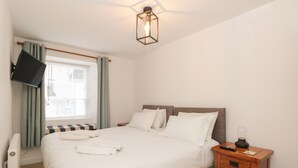 A neatly made bed with white linens, a wooden nightstand with drawers, a wall-mounted TV, and a window with curtains.