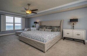 Une chambre avec un grand lit, un ventilateur de plafond, une table de chevet, une lampe et une vue sur l’océan.