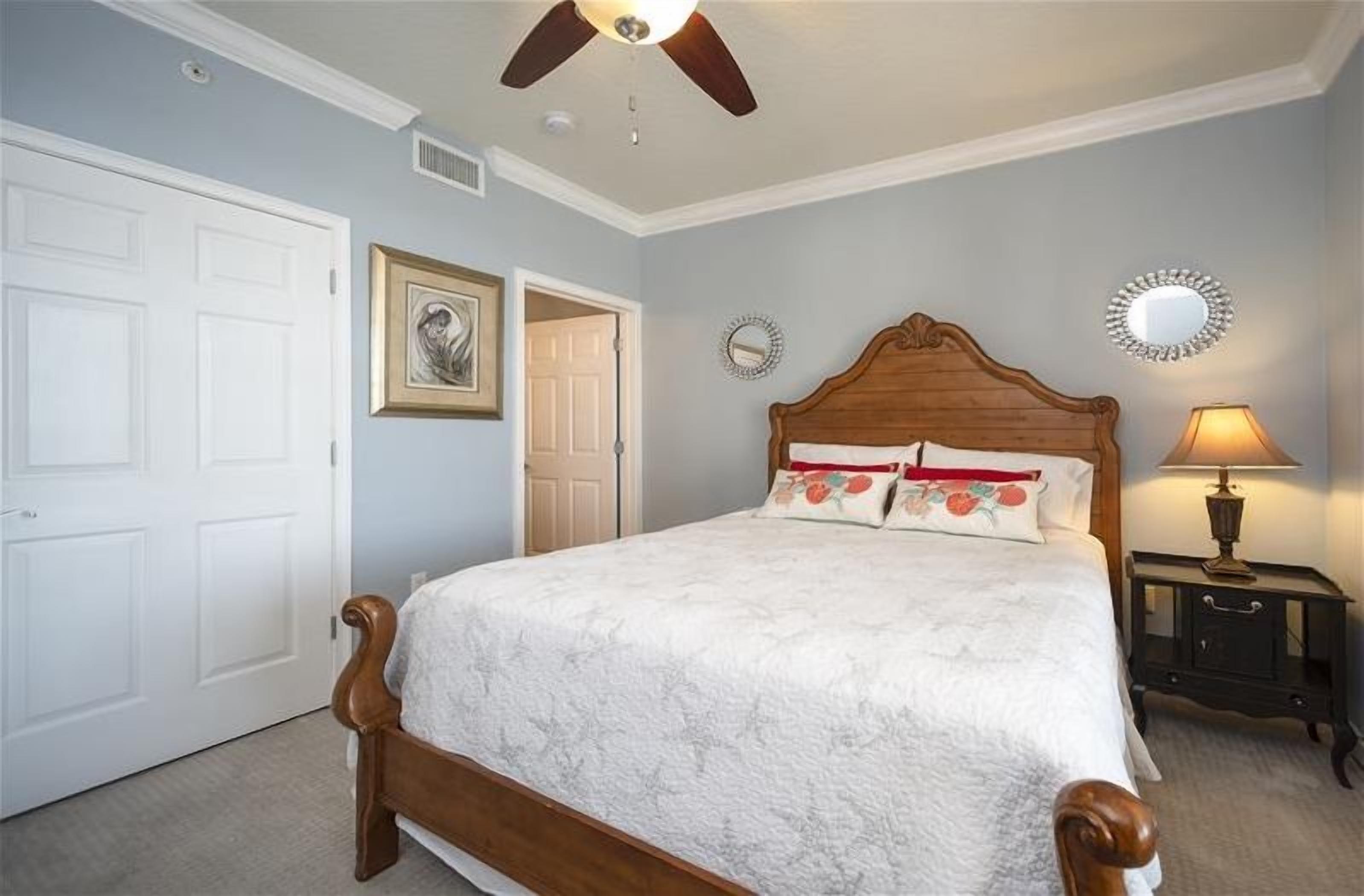A bedroom with a wooden bed, white walls, a ceiling fan, a bedside table with a lamp, and a door.