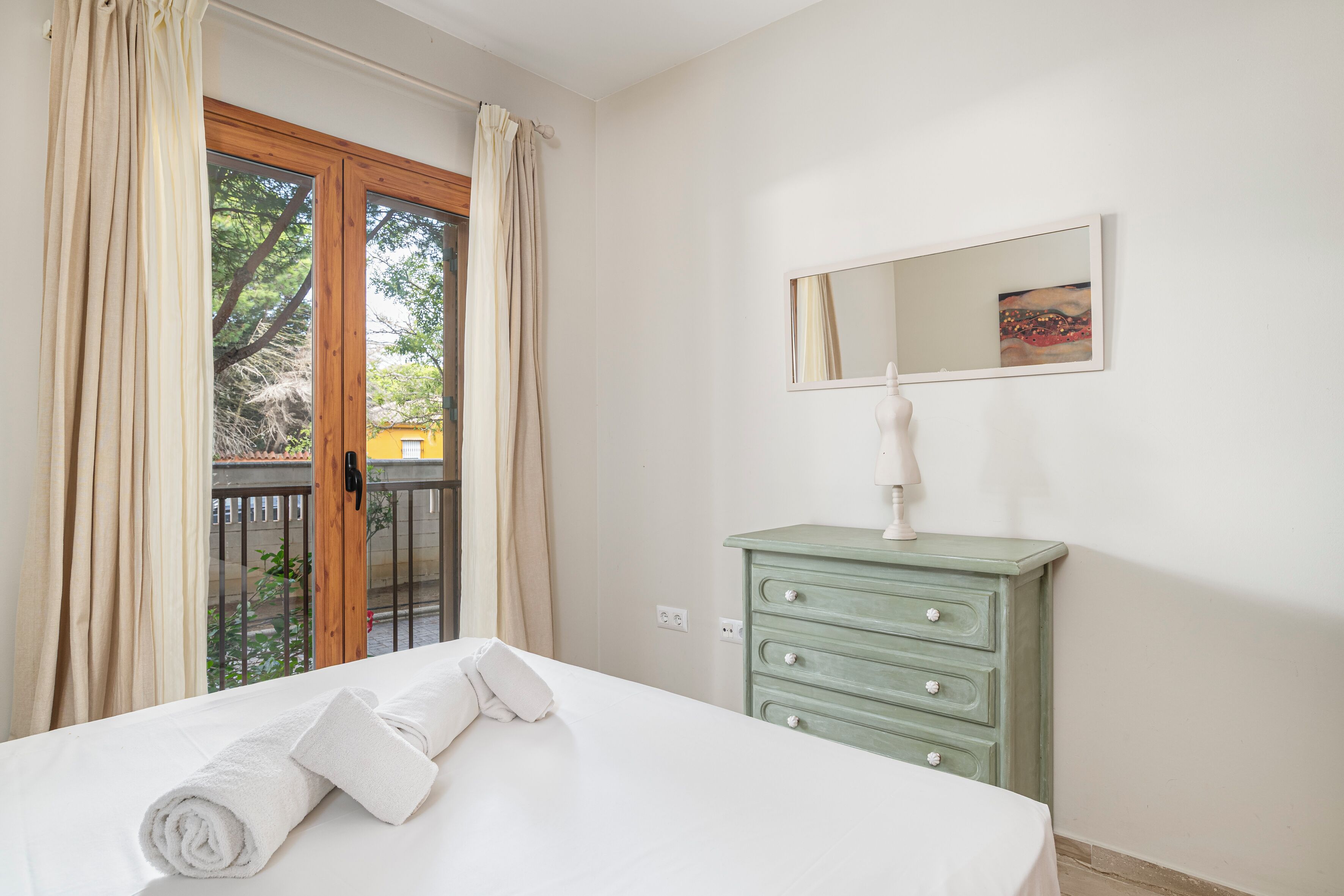 A bedroom with a bed, a dresser, a mirror, and a view of the outside.