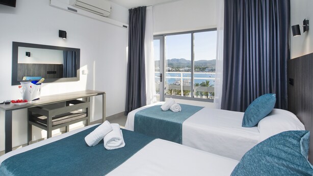 Une chambre d’hôtel avec deux lits, un bureau, une grande fenêtre avec des rideaux, une vue sur la mer et un téléviseur fixé au mur.