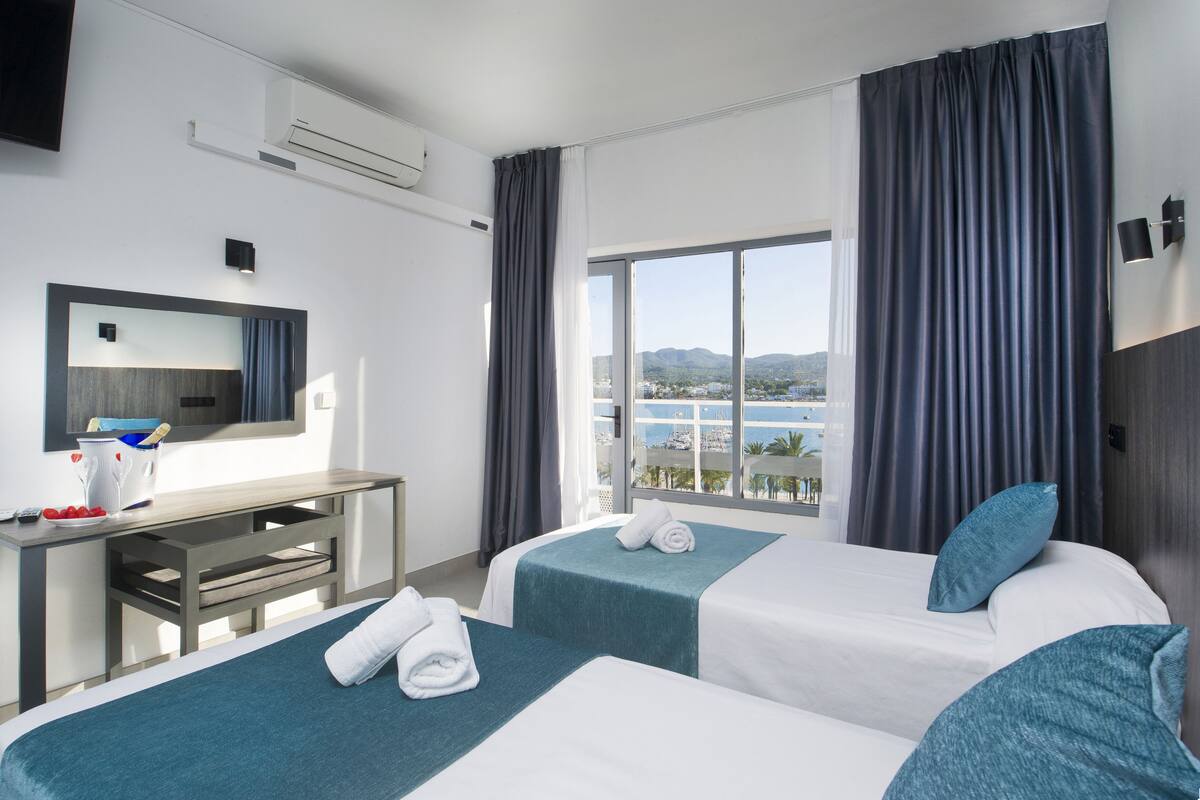 Ein Hotelzimmer mit zwei Betten, einem Schreibtisch, einem großen Fenster mit Vorhängen und Meerblick sowie einem Wand-Fernseher.