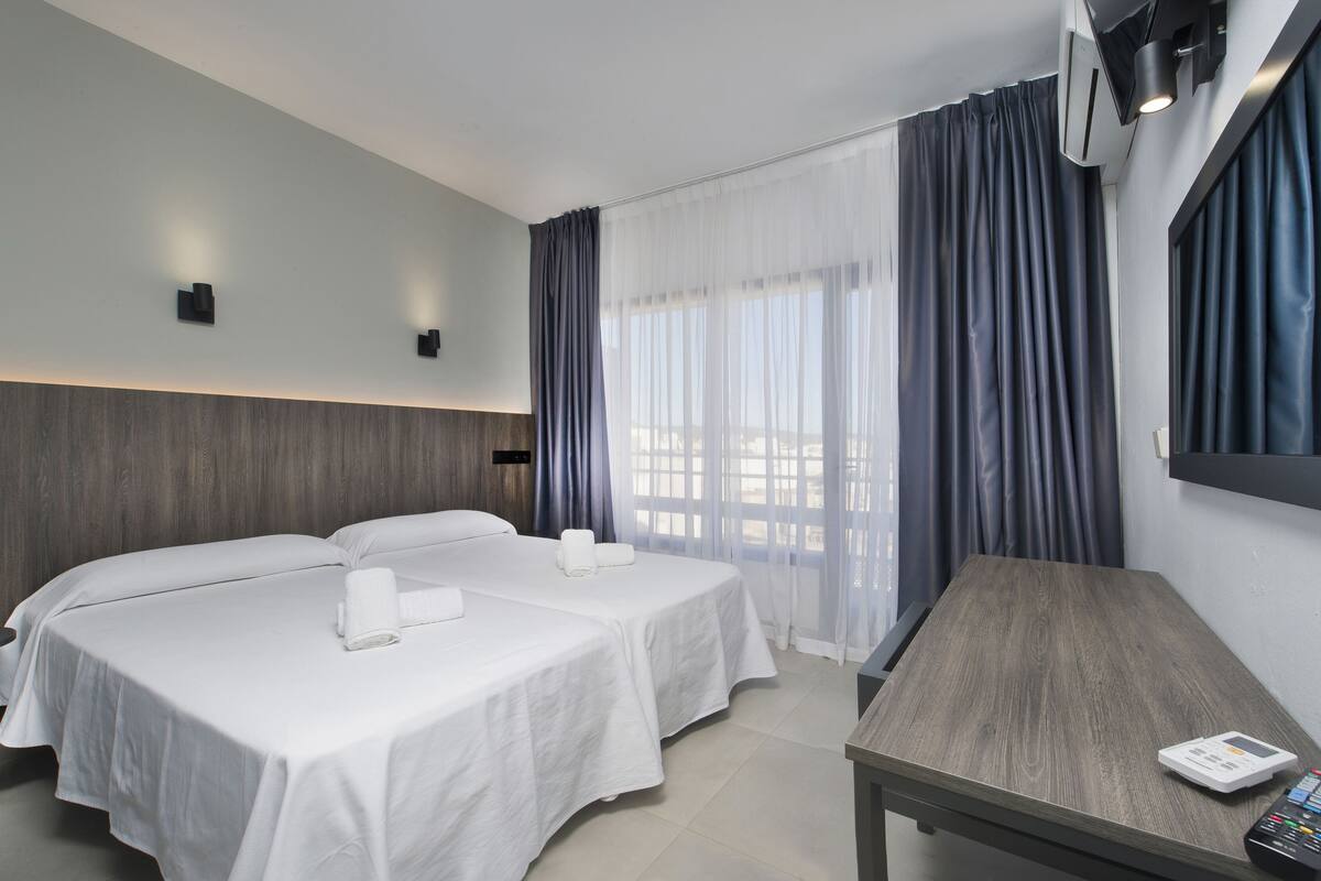 Ein Hotelzimmer mit zwei Betten, einem kleinen Tisch, einem Fernseher und einem Fenster mit Vorhängen.