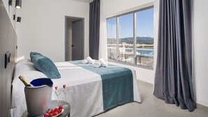 Une chambre d’hôtel avec un lit, une petite table sur laquelle se trouve un seau à champagne et des fraises, une vue sur la mer et les montagnes à travers de grandes fenêtres, et un balcon avec une chaise.