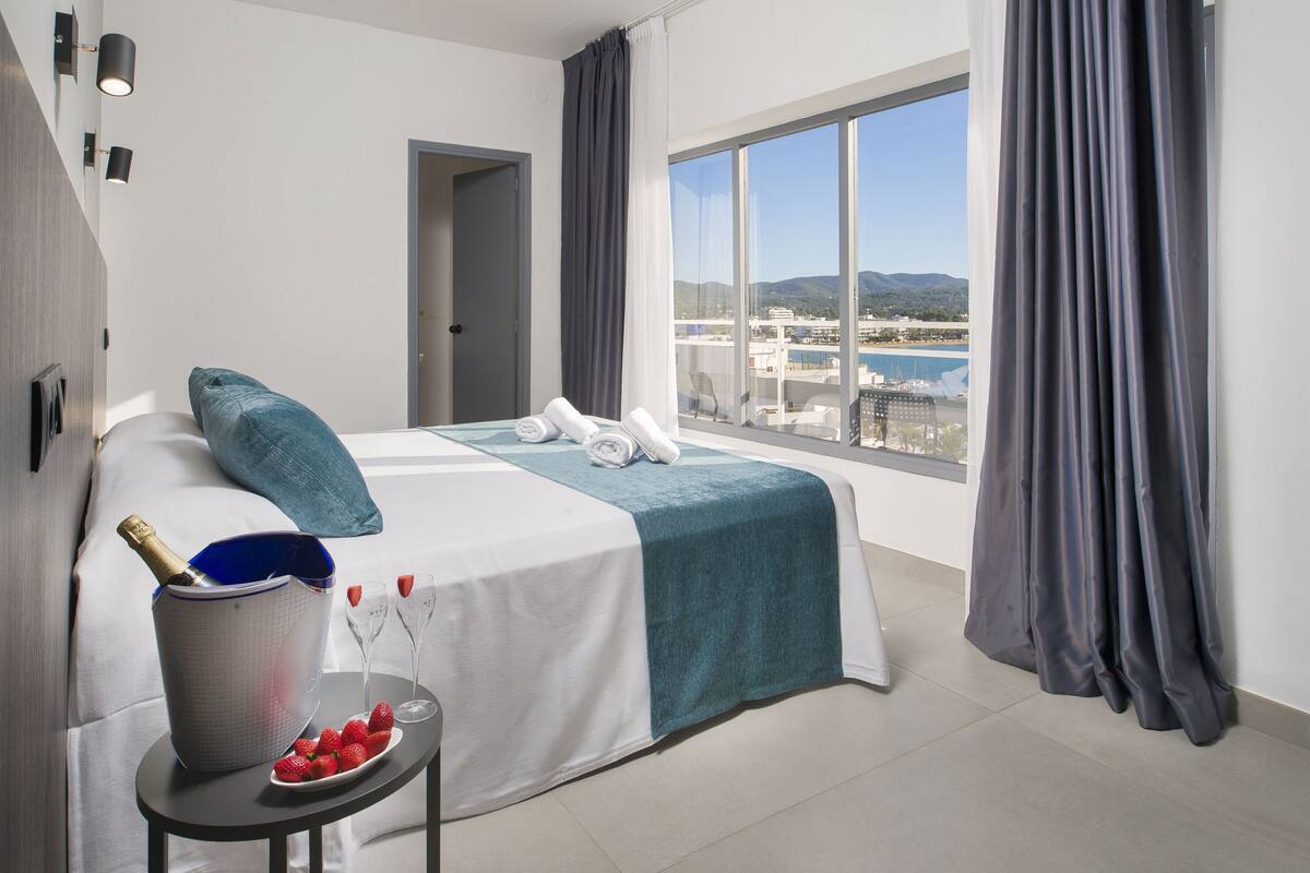 Ein Hotelzimmer mit Bett, einem kleinen Tisch mit Eimer für Champagner und Erdbeeren, Meer- und Bergblick durch große Fenster sowie ein Balkon mit Stuhl.