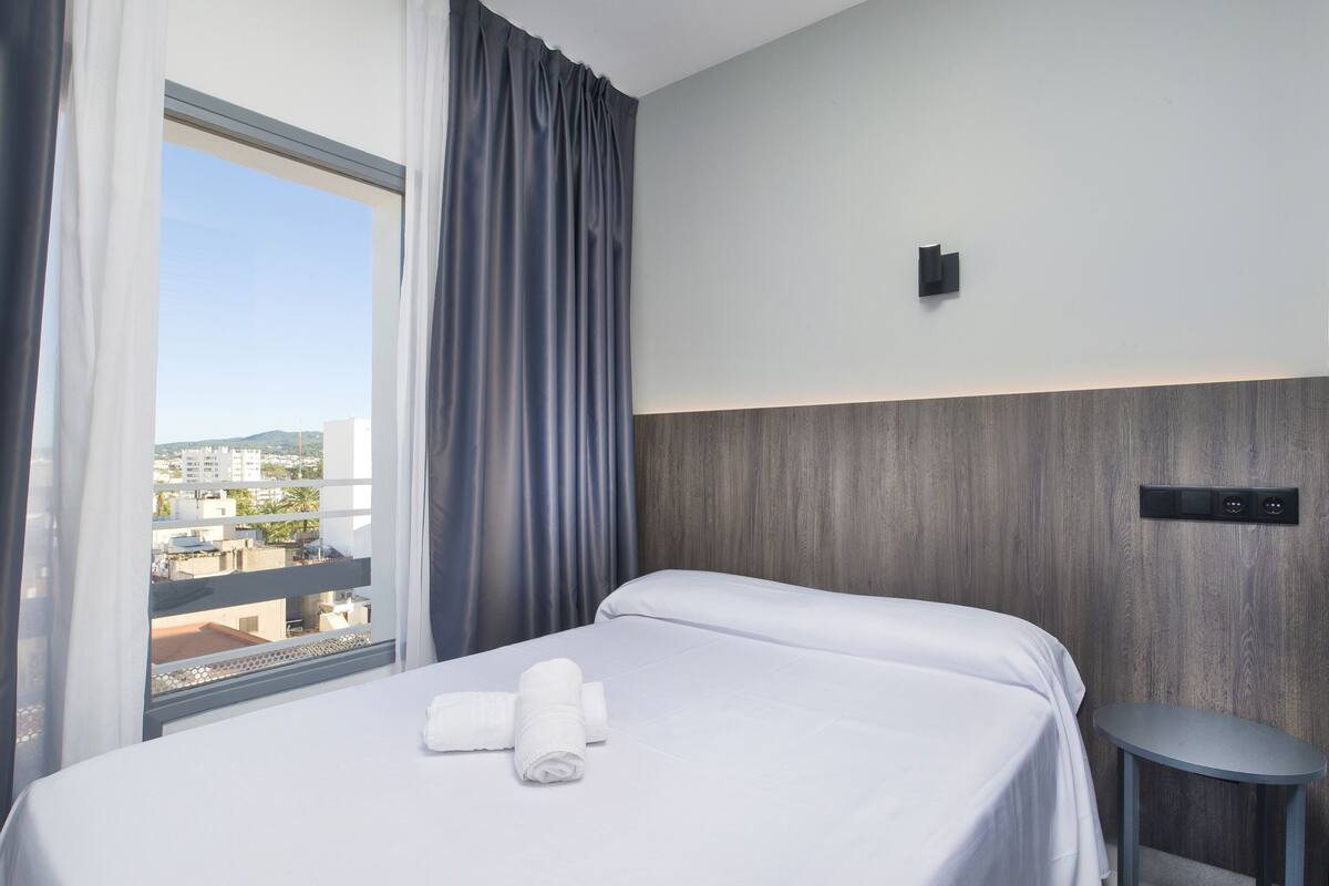 Ein Hotelzimmer mit Bett, kleinem Tisch, einem Fenster mit Vorhängen und Blick auf Gebäude.