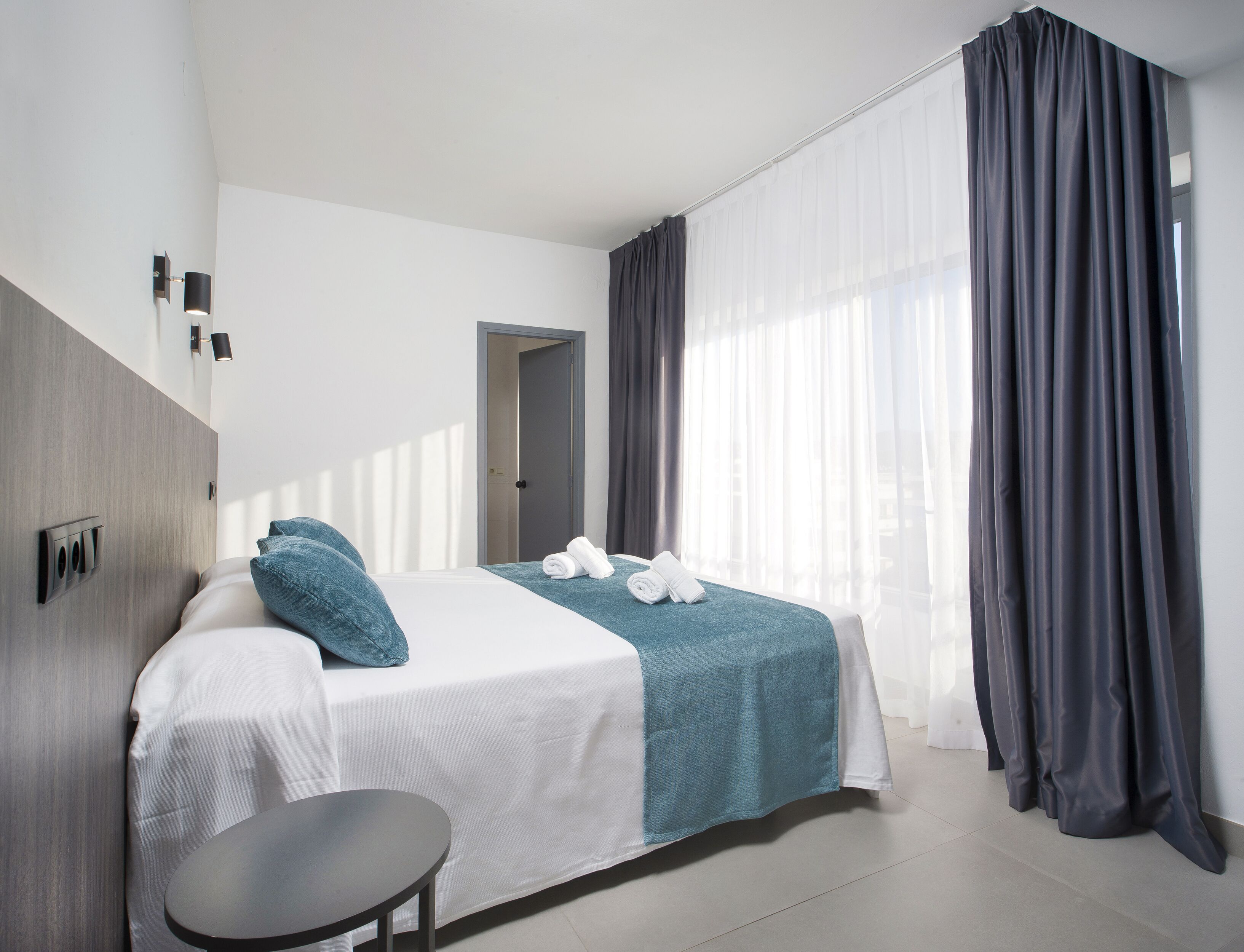 A modern hotel room with a bed, a small table, and a door leading to another room.