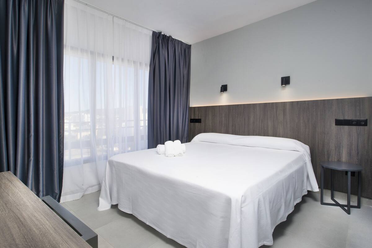 Ein modernes Hotelzimmer mit einem großen Bett, einem Nachttisch und Blick durch transparente Vorhänge.
