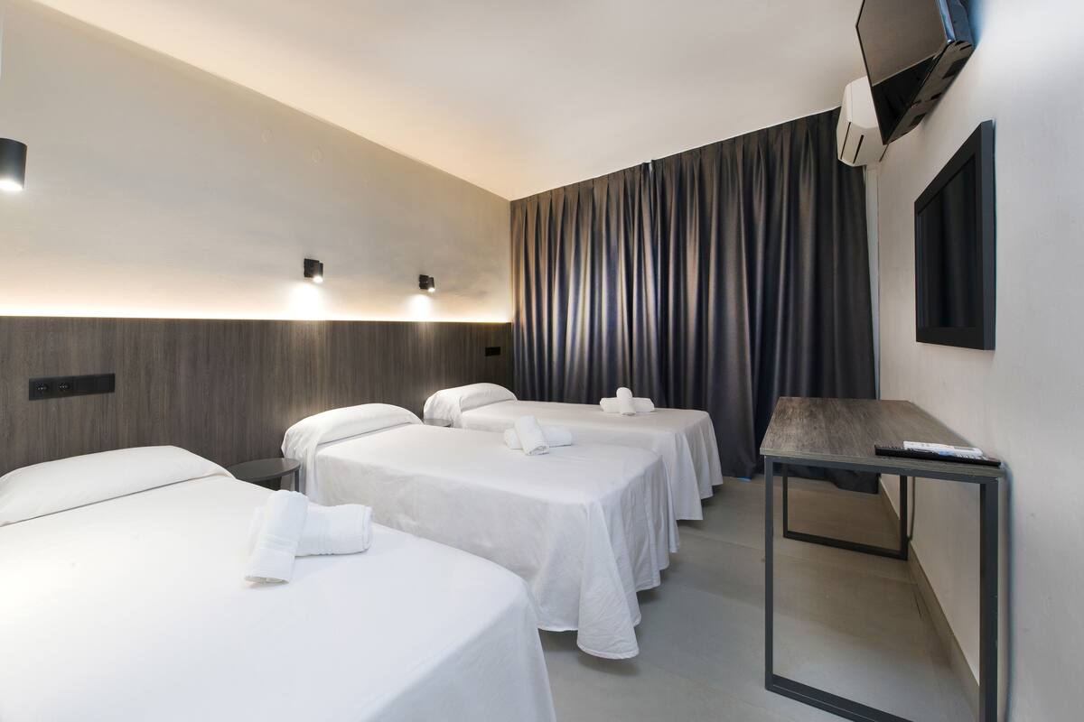 Ein Hotelzimmer mit drei Einzelbetten, einem kleinen Tisch, einem Fernseher und einem Vorhang.
