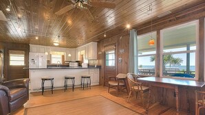 Une cuisine avec des meubles en bois, un réfrigérateur, des tabourets de bar et une vue sur l’océan grâce à de grandes fenêtres.