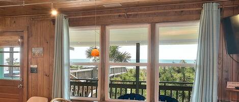 A wooden interior with a wooden table and chairs, large windows with a view of the beach and palm trees, and a television on the wall.