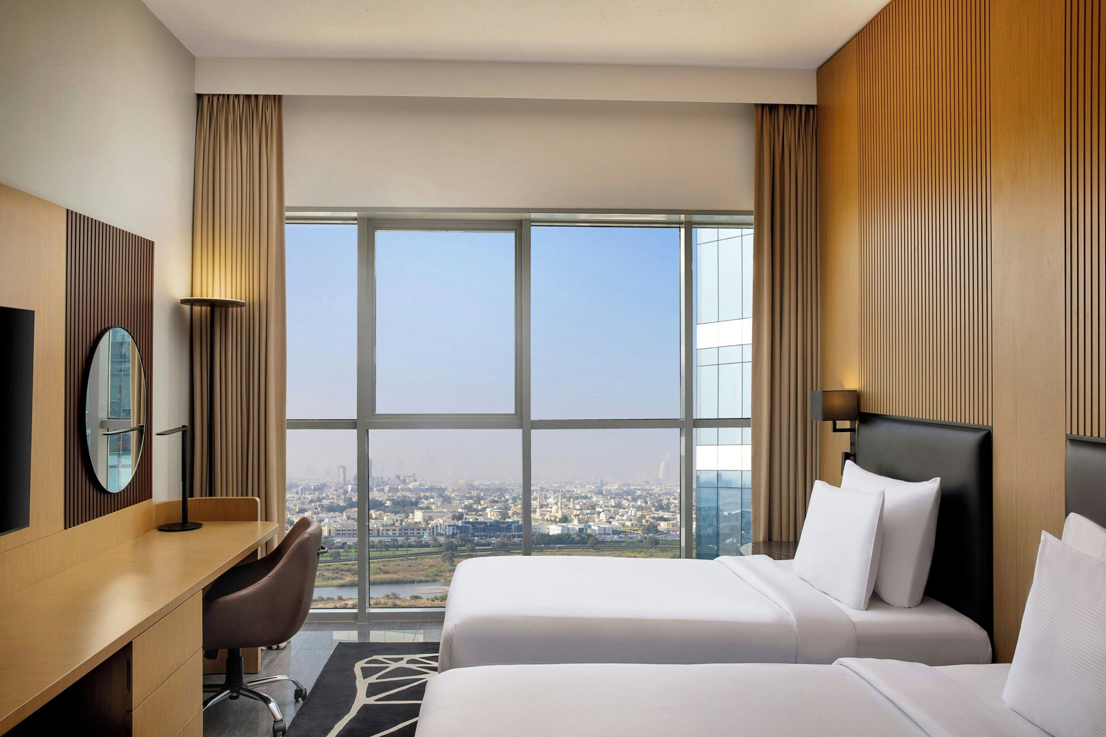 A hotel room with two beds, a desk, a chair, a TV, and a large window offering a city view.