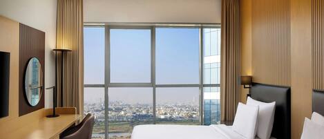 A hotel room with two beds, a desk, a chair, a TV, and a large window offering a city view.